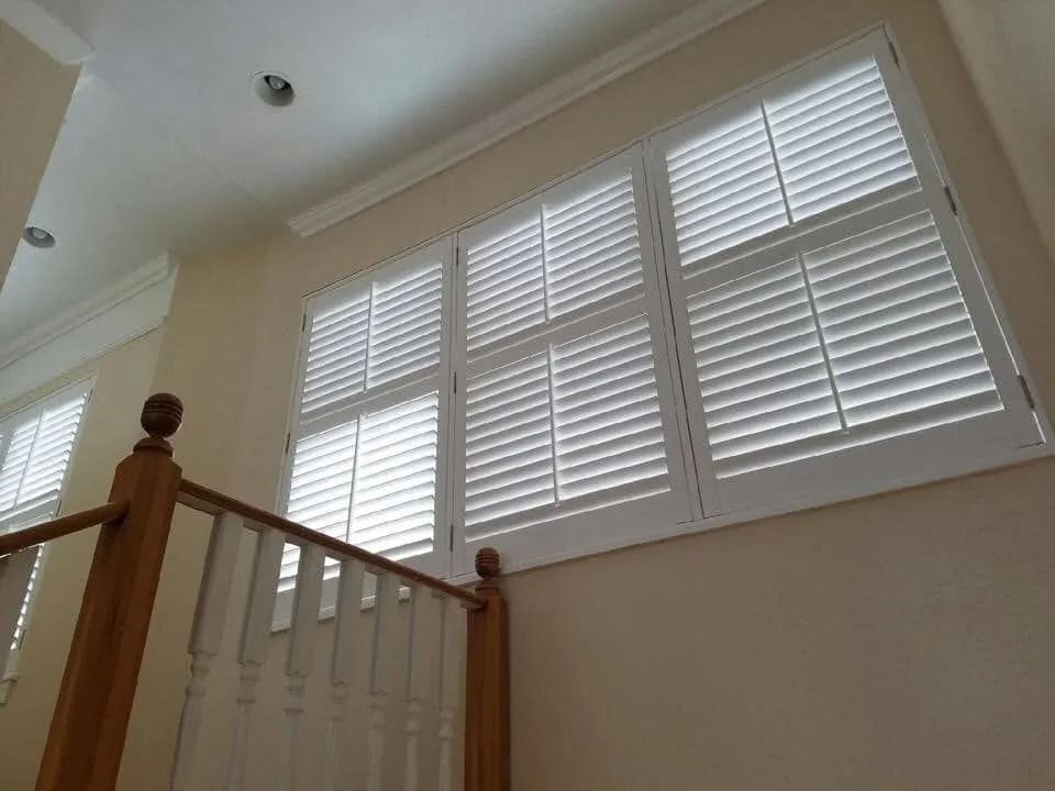Interior staircase with wooden railing beside large white plantation shutters and beige walls