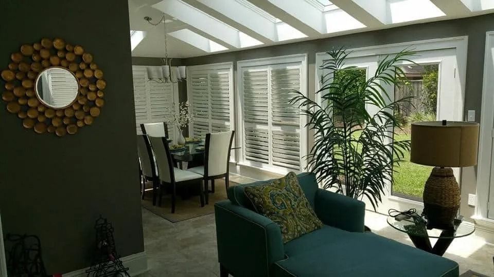 Bright living room with green sofa, tall plants, skylight ceiling, and dining area by white shutters