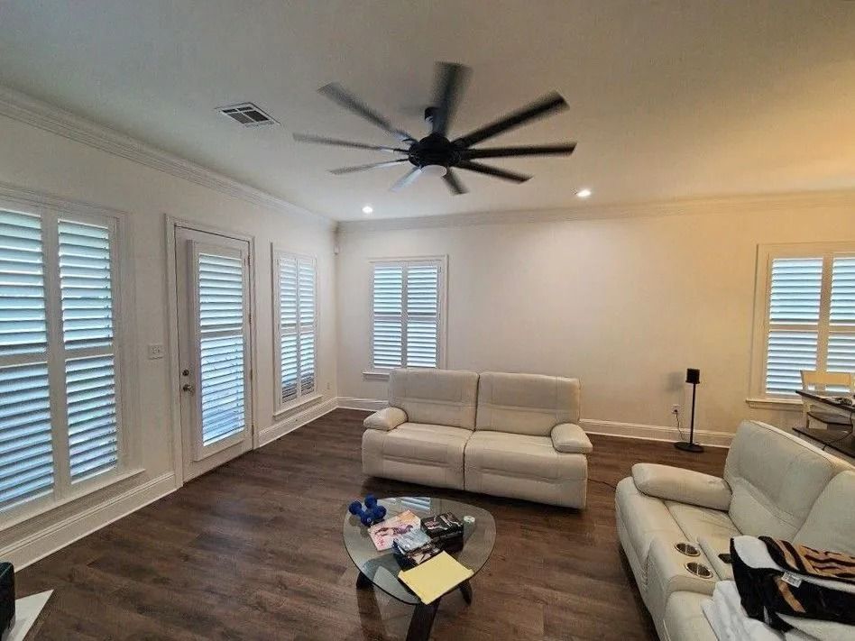 Bright living room with white sofas, ceiling fan, wood floors, and plantation shutters.