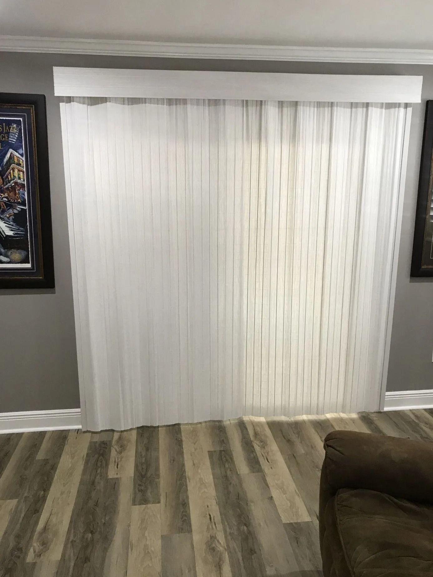 Living room with vertical white blinds over a sliding glass door, gray wood-look floor, and a brown sofa edge