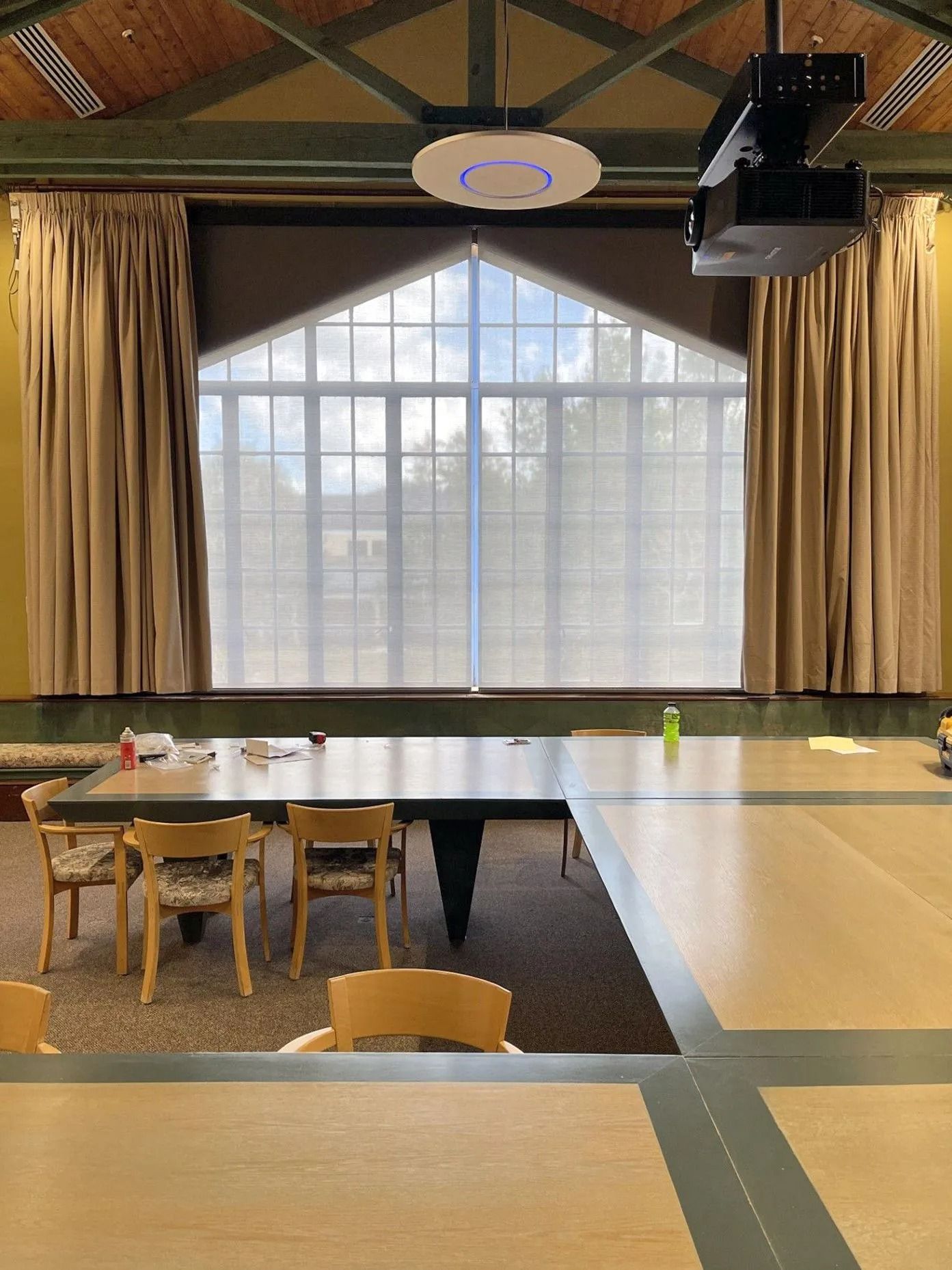 Empty classroom with tables, chairs, large curtained windows, and a ceiling projector.