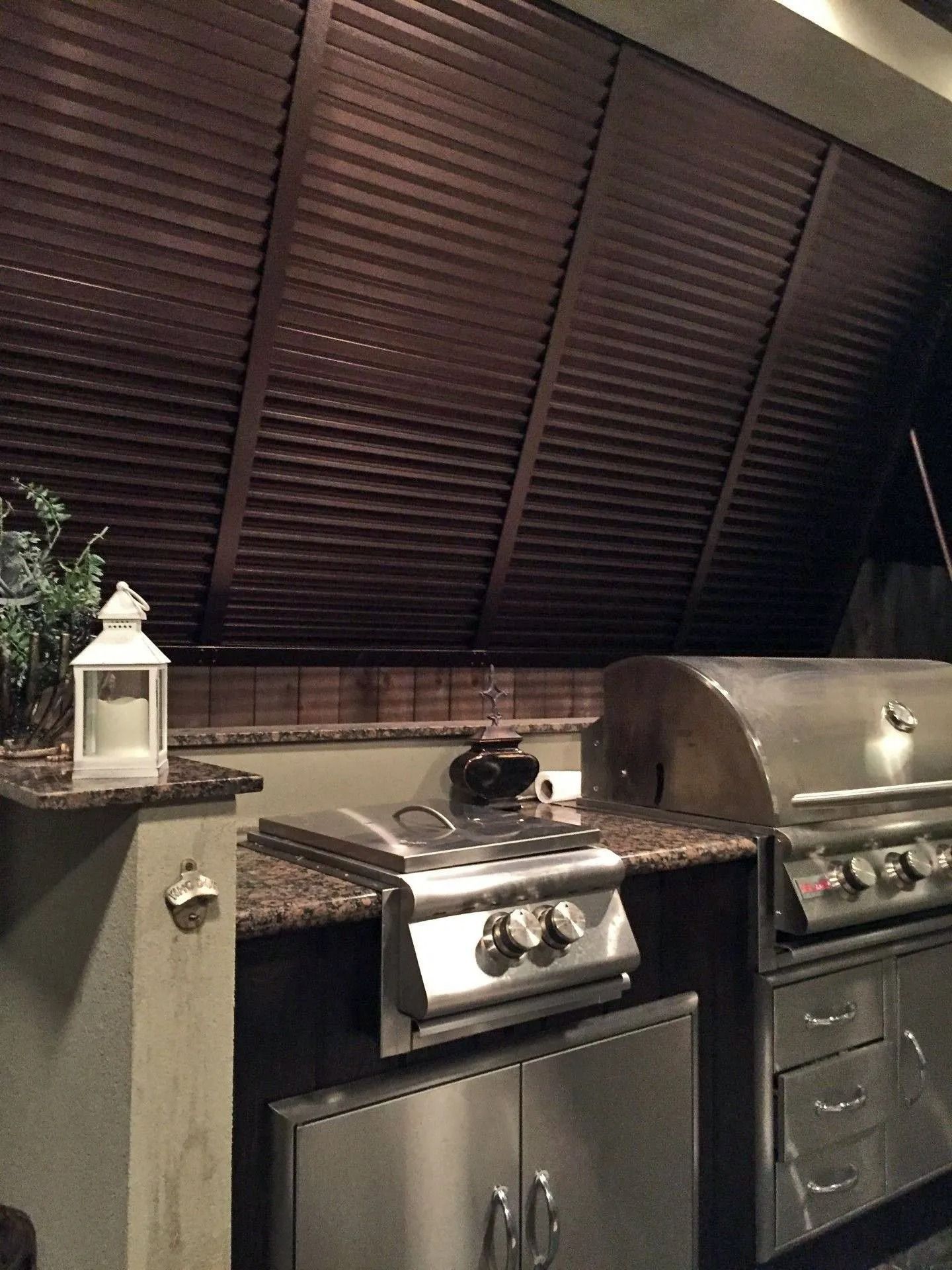 Outdoor kitchen with grill, stainless steel cabinets, and a lantern under a slatted roof.