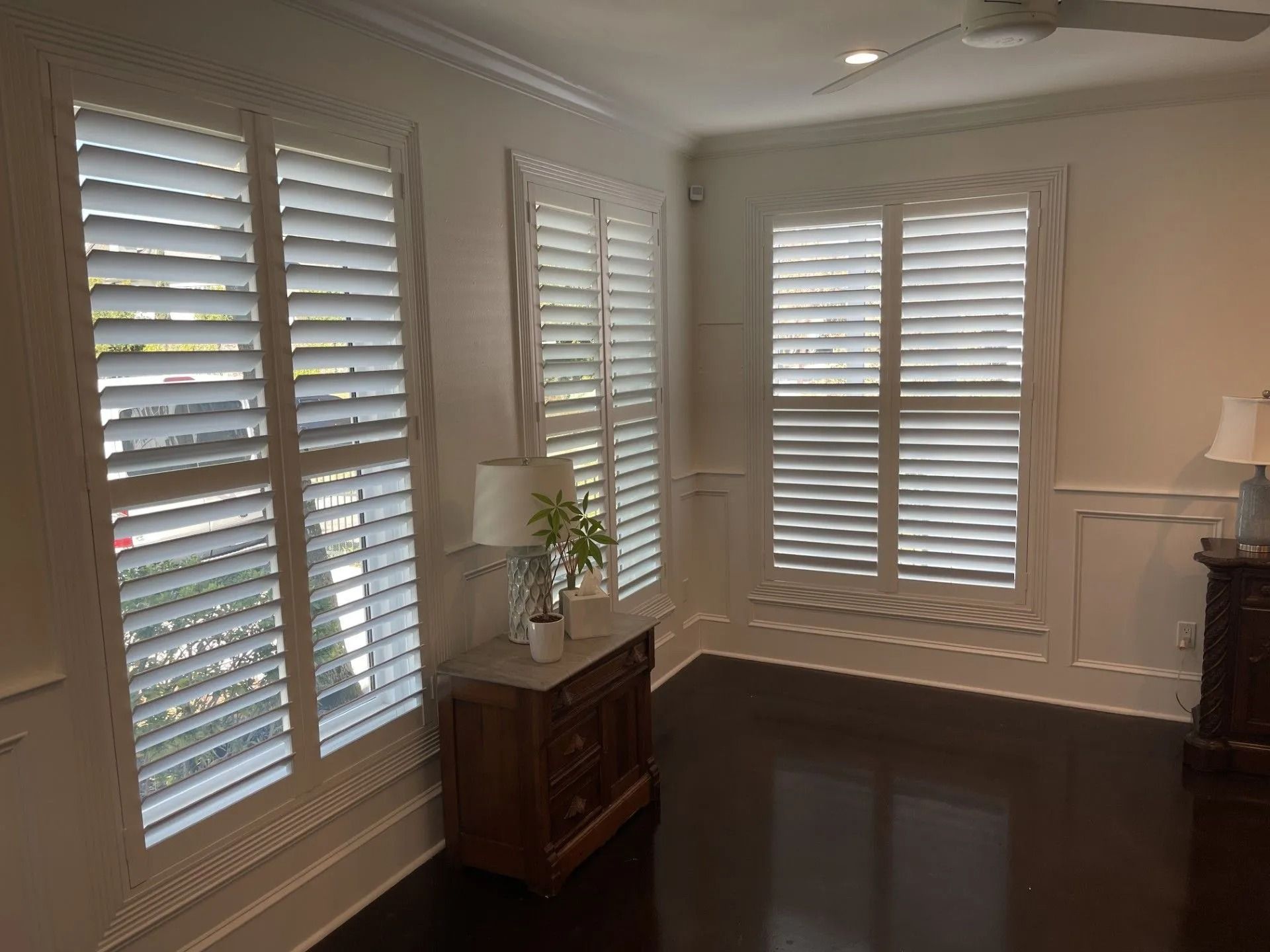 Bright room with white plantation shutters, dark hardwood floors, and a small wooden cabinet with a plant.