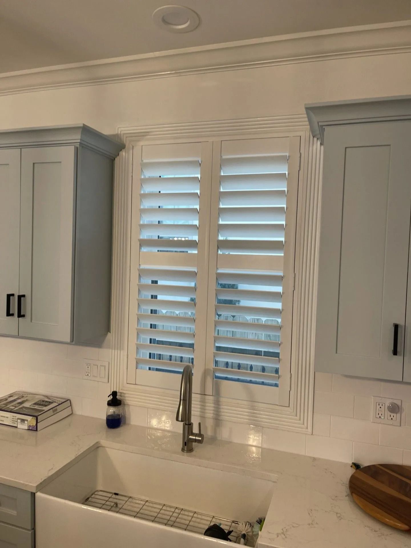 White kitchen sink beneath shuttered window with gray cabinets and chrome faucet