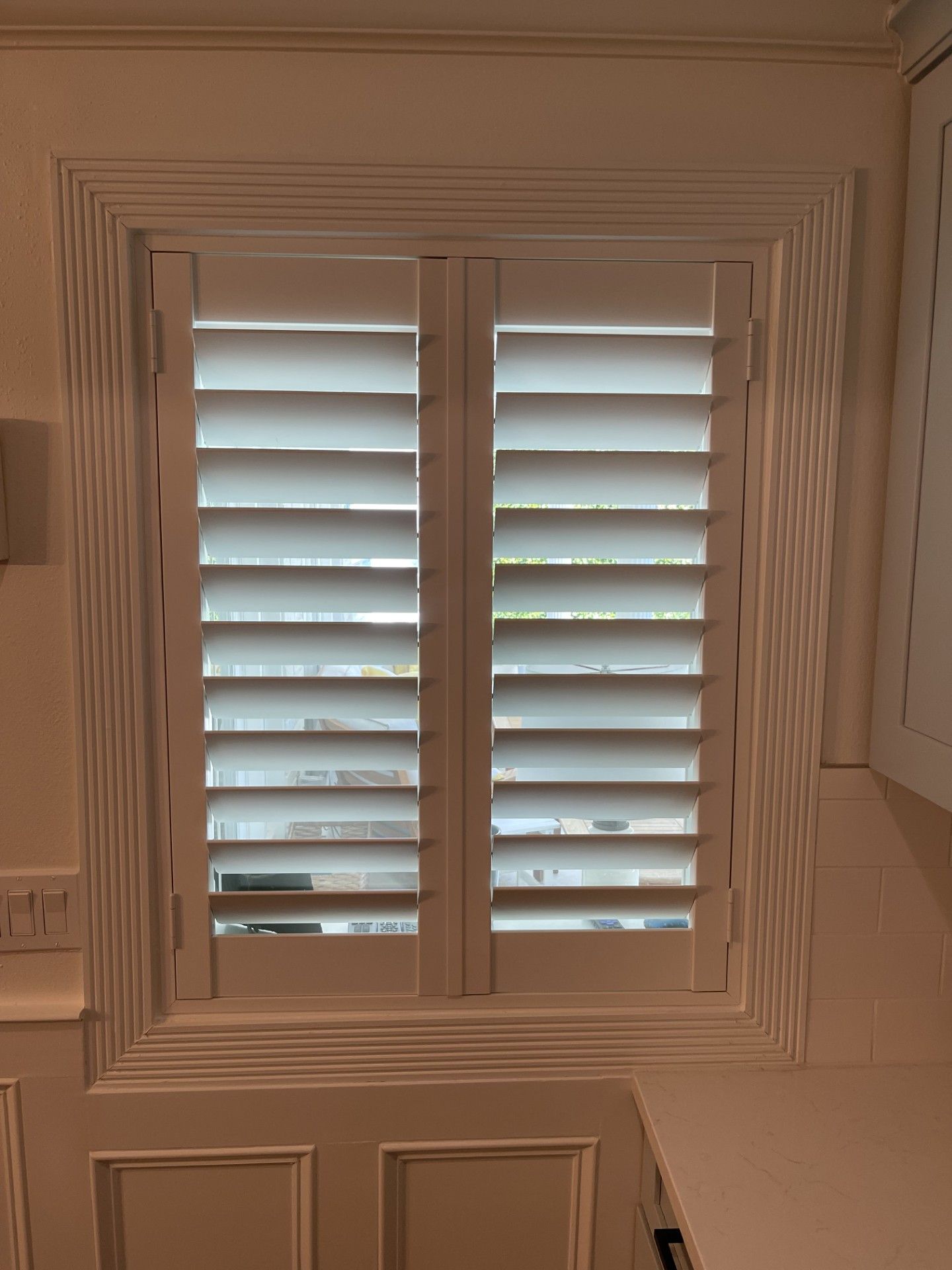 White shuttered window with slatted blinds, framed by beige trim and a wall panel below.