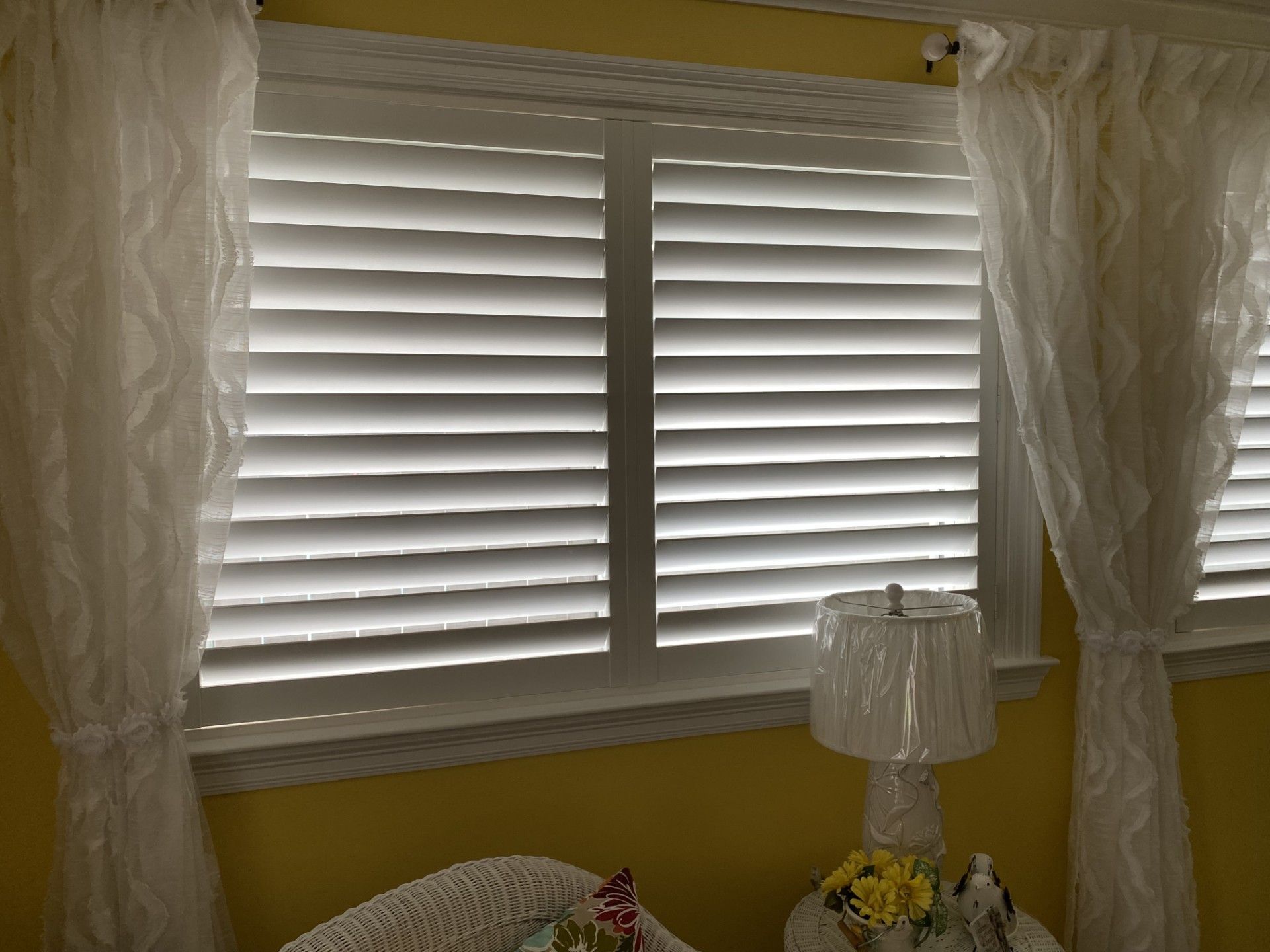 Bedroom window with white shutters, lace curtains, and a small lamp on a bedside table