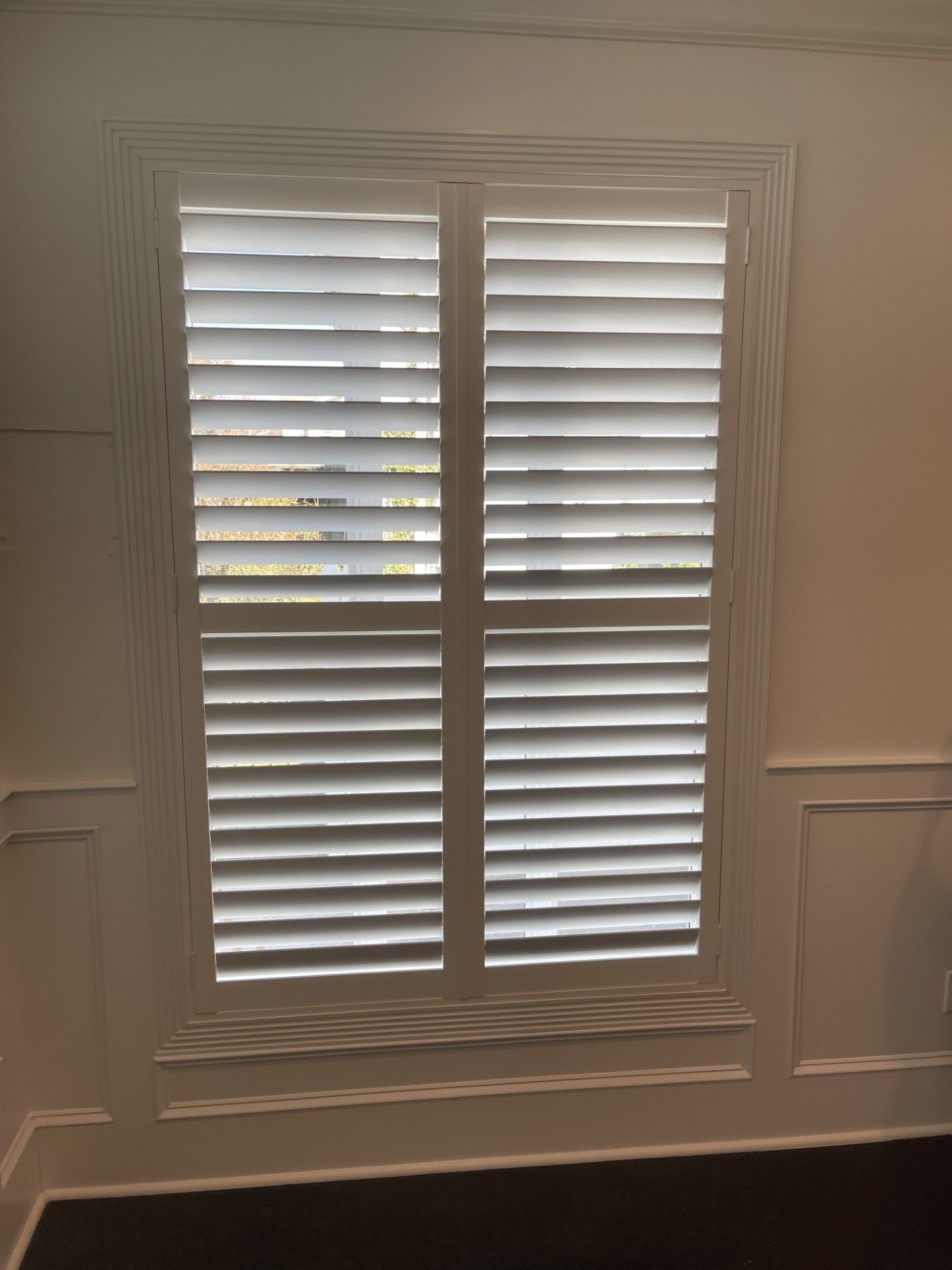 White plantation shutters on a window in a beige interior wall above dark hardwood flooring