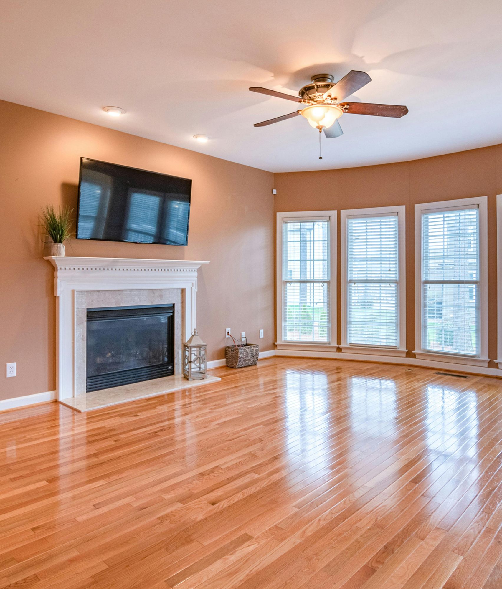 Living room with fireplace, TV, and three windows, with shiny hardwood floors and a ceiling fan.