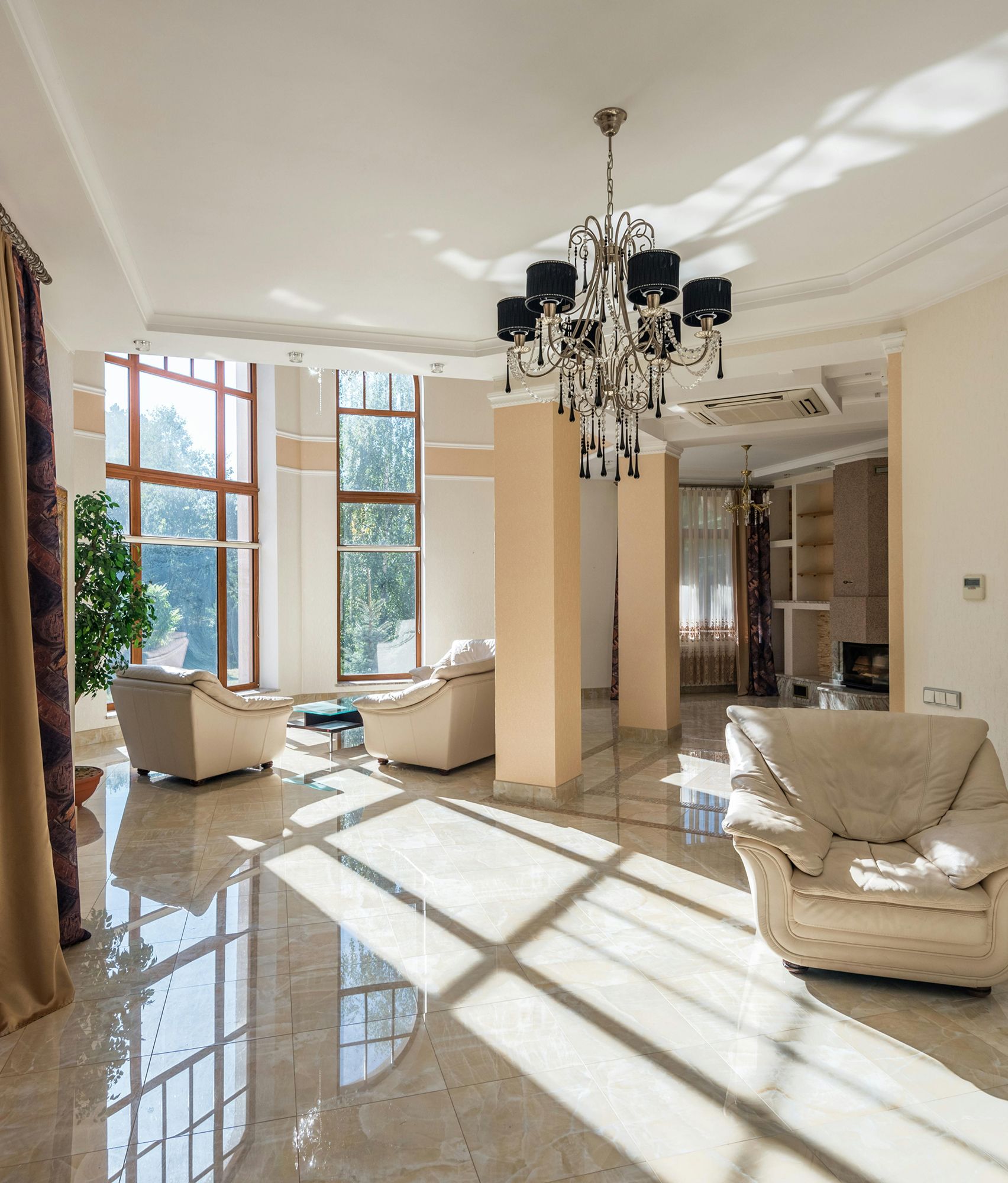 Luxurious living room with chandelier, cream-colored armchairs, large windows, and glossy tiled floor.