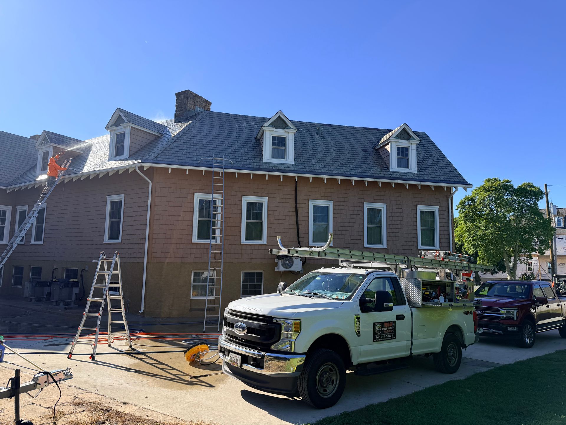 Workers on ladders beside a brown building and white pickup truck, likely roofing or construction work. Sunny day.
