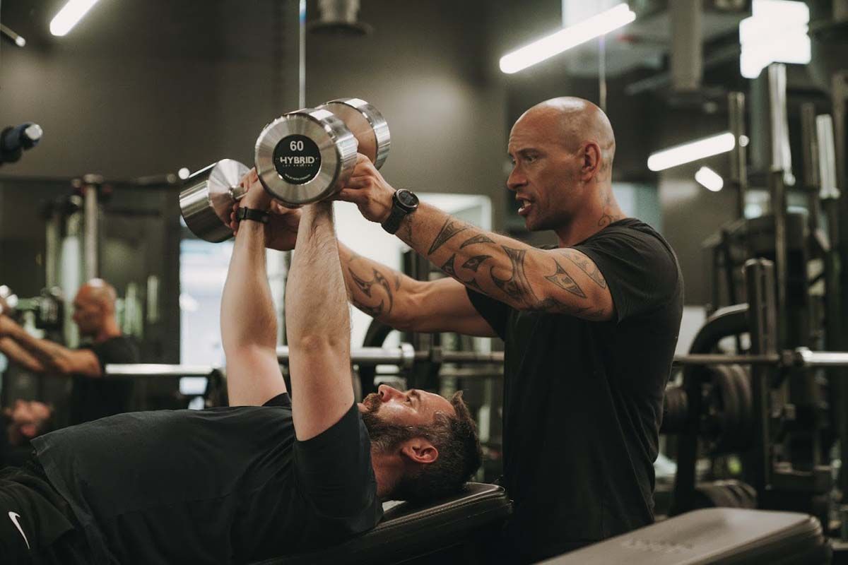 A man is lifting a dumbbell while another man watches in a gym.