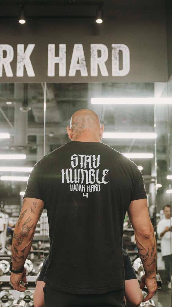 A man is standing in a gym wearing a black t-shirt that says `` stay humble ''.