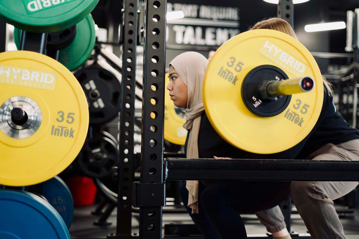 A woman in a hijab is squatting with a barbell in a gym.