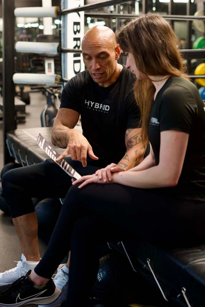 A man and a woman are sitting on a bench in a gym looking at a tablet.