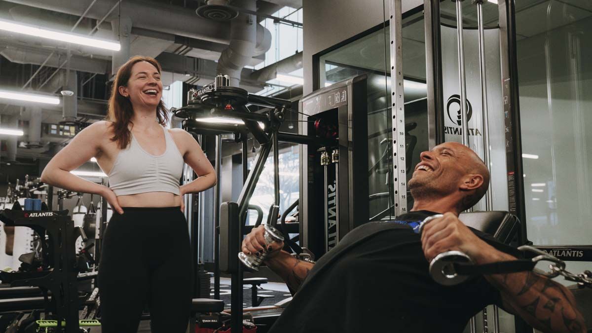 A woman is standing next to a man lifting weights in a gym.