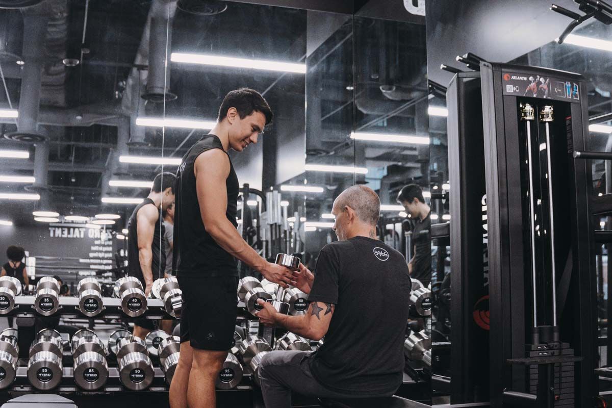 A man is talking to another man in a gym while holding a dumbbell.