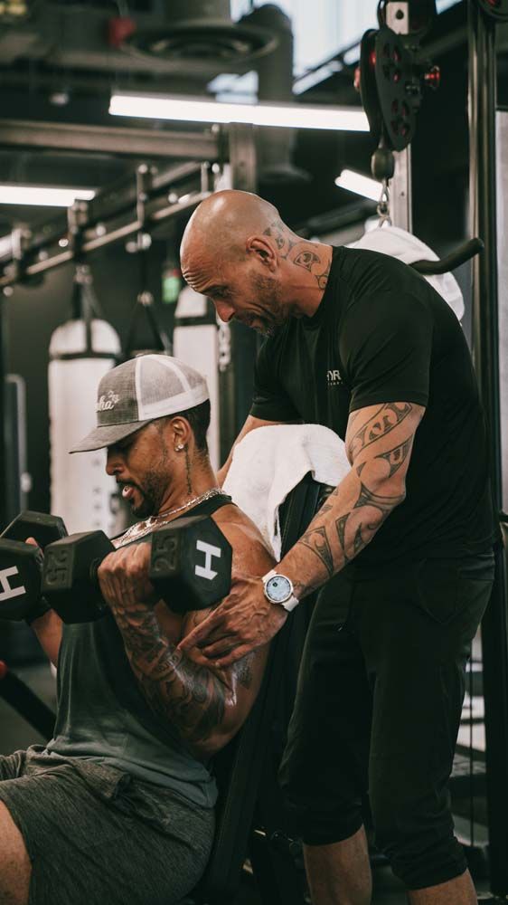 A man is helping another man lift a dumbbell in a gym.