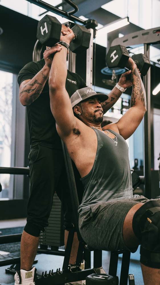 A man is lifting dumbbells while another man watches in a gym.