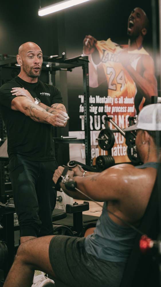 A man is standing next to a man sitting on a machine in a gym.
