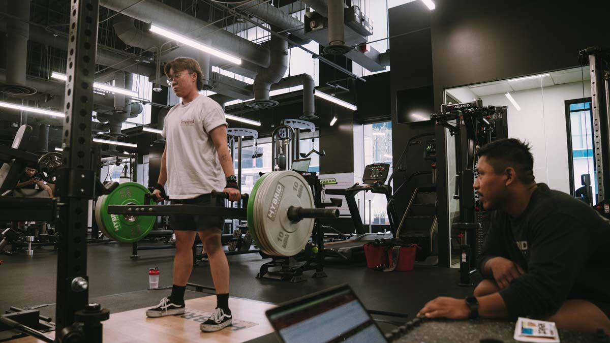 A man is lifting a barbell in a gym while another man looks on.