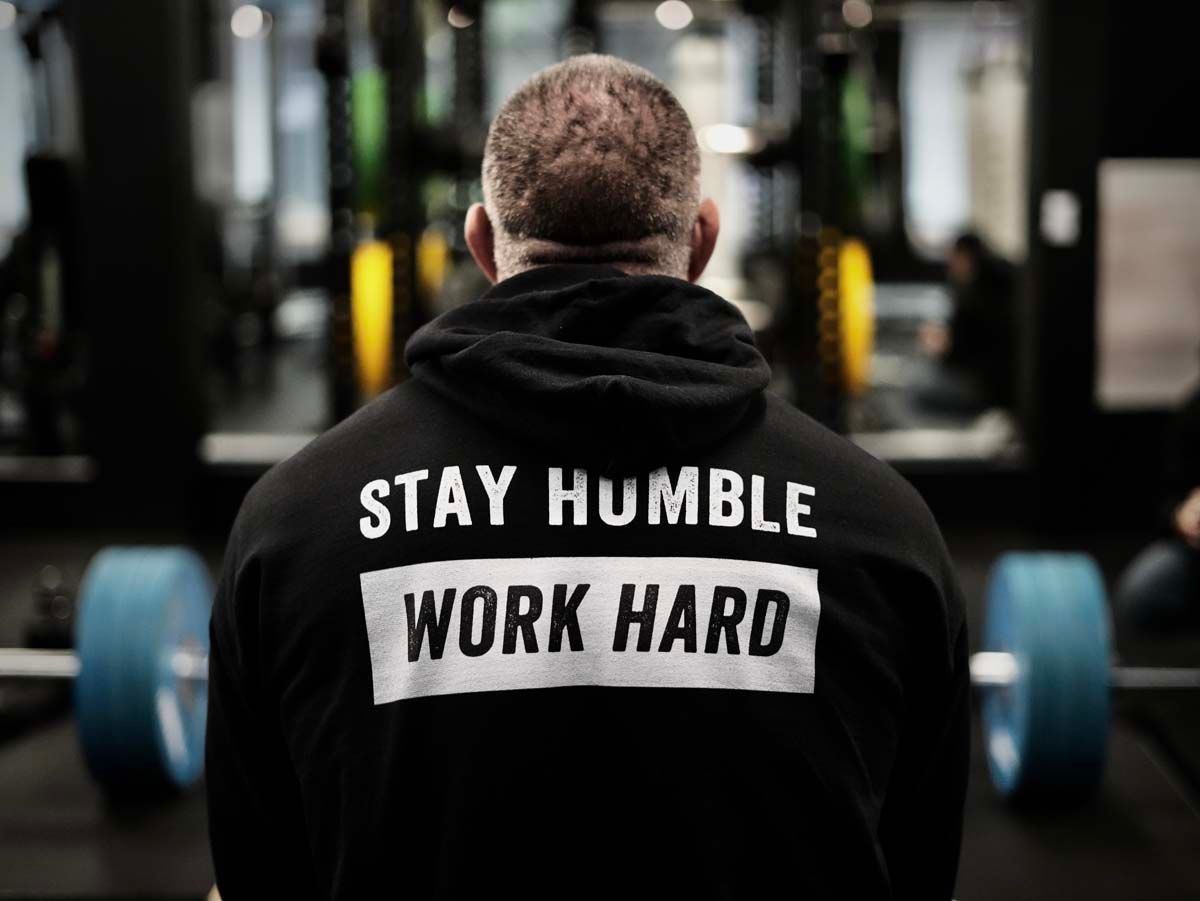A man wearing a hoodie that says `` stay humble work hard '' is standing in a gym.