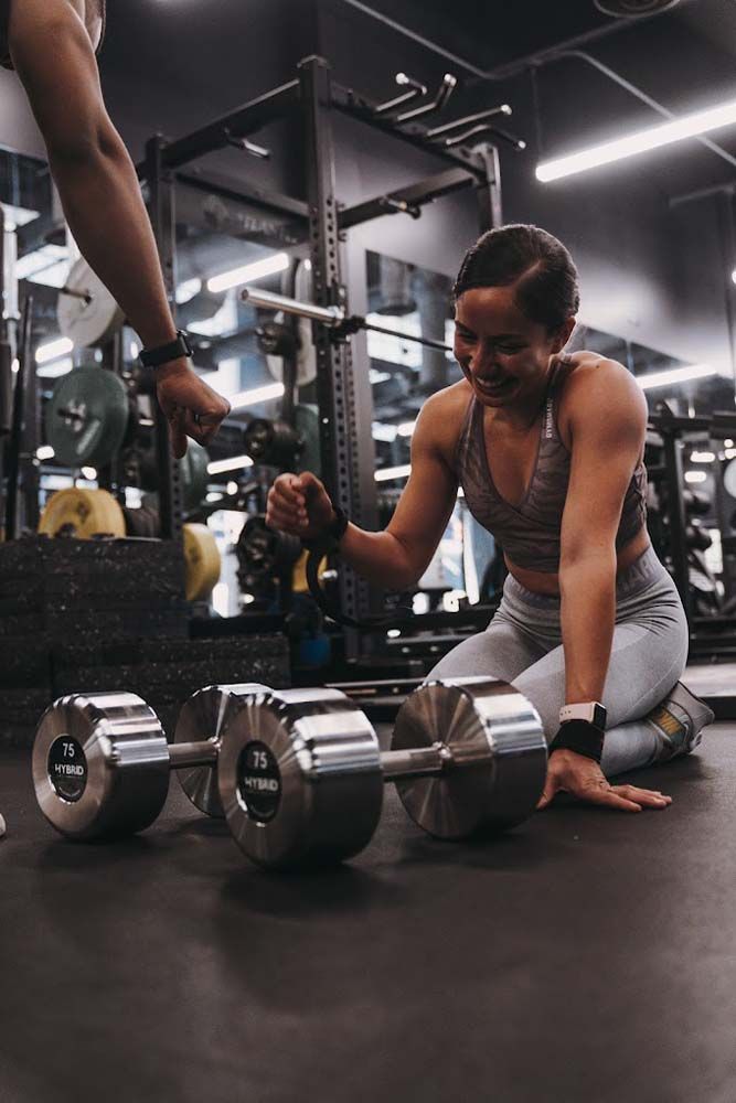 A woman is kneeling down next to a pair of dumbbells in a gym.