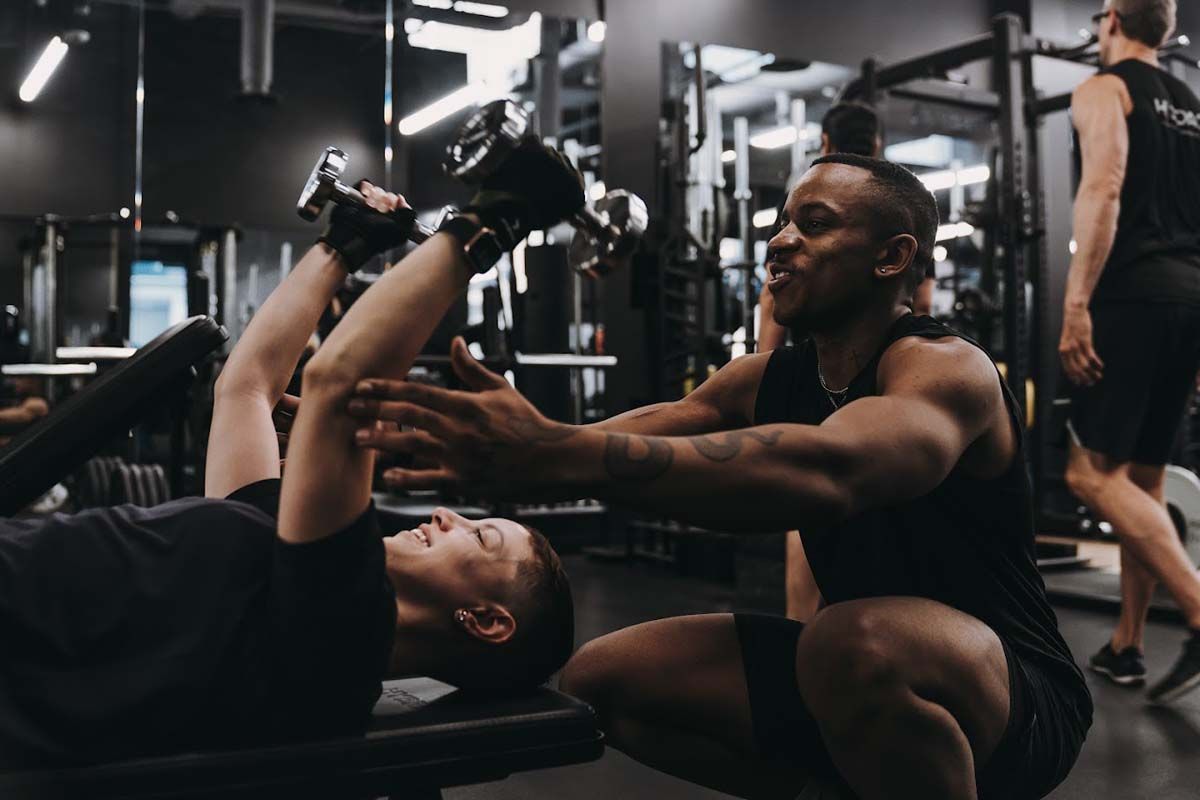 A man is helping another man lift dumbbells in a gym.