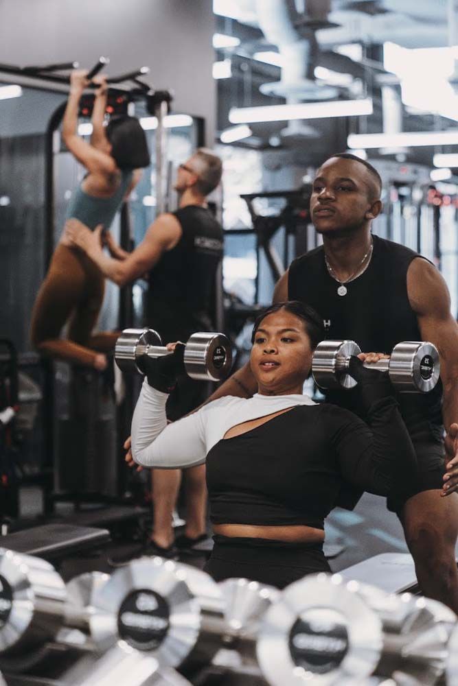 A man is helping a woman lift dumbbells in a gym.