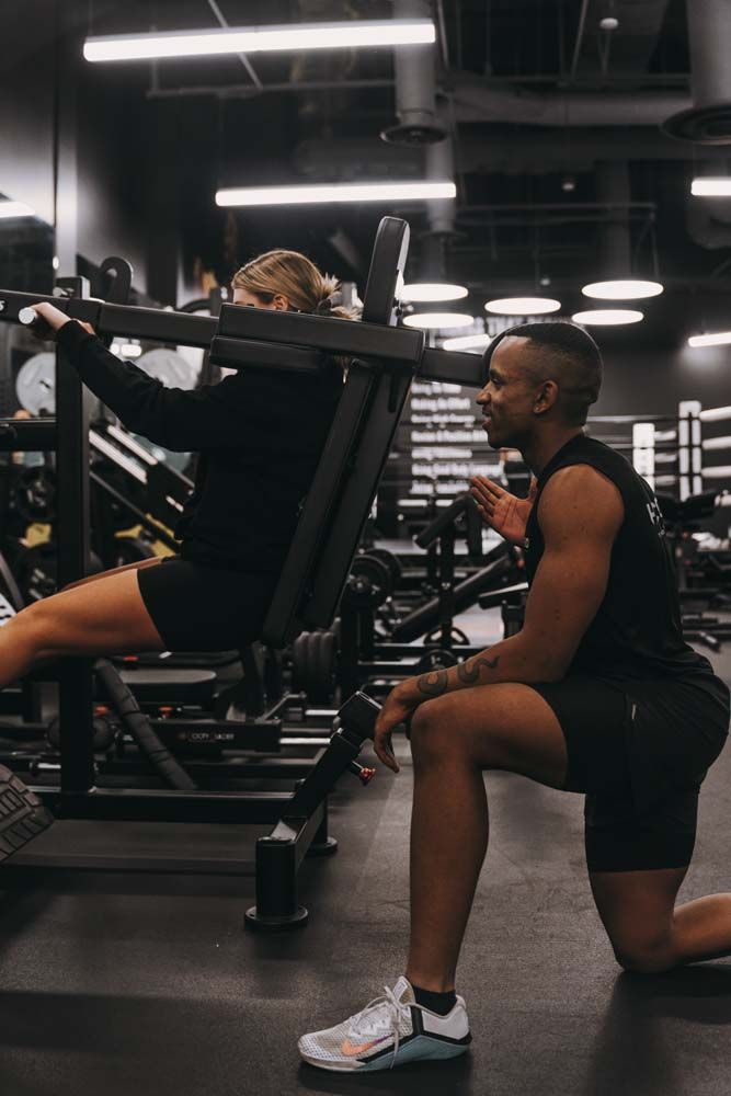 A man is squatting next to a woman using a machine in a gym.