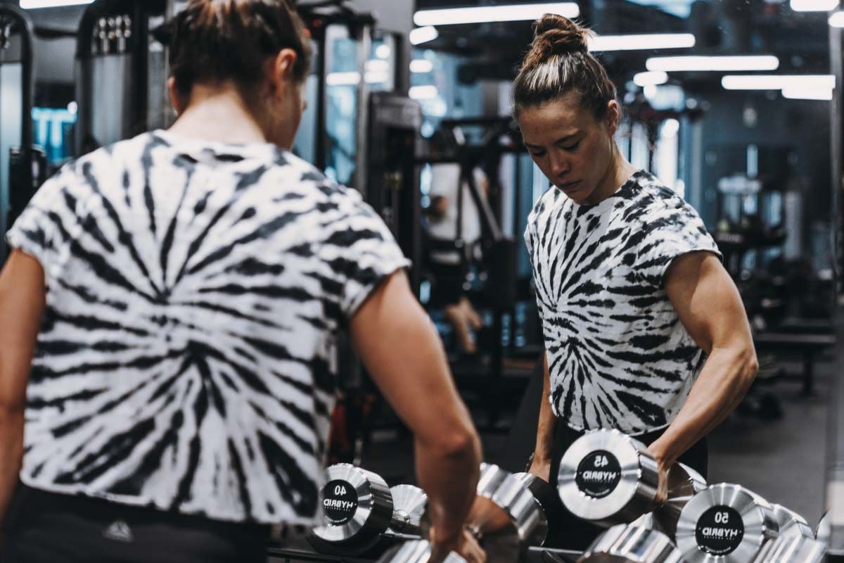 A woman is lifting a dumbbell in front of a mirror in a gym.