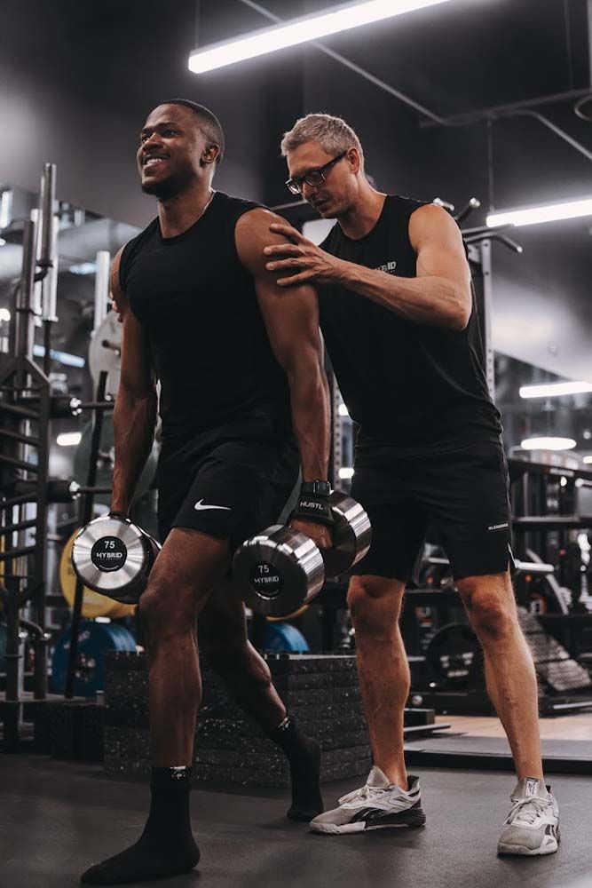 A man is helping another man lift dumbbells in a gym.
