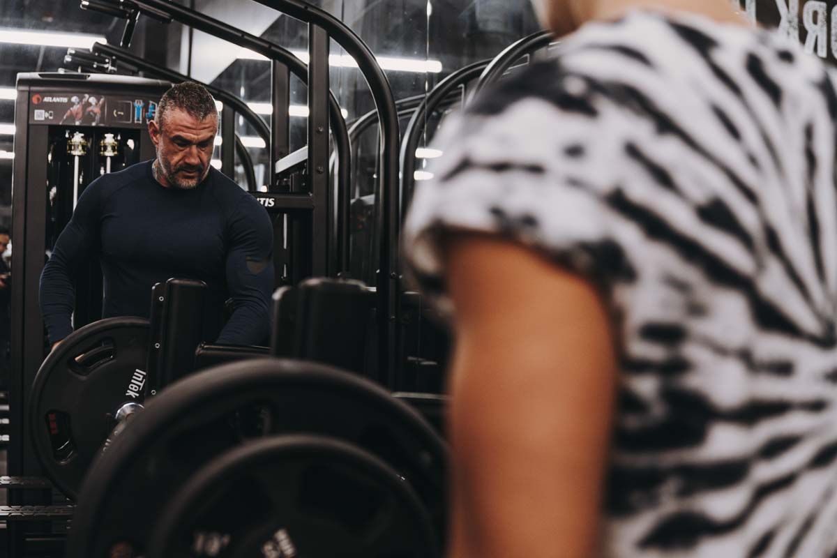 A man is lifting weights in a gym while a woman looks on.