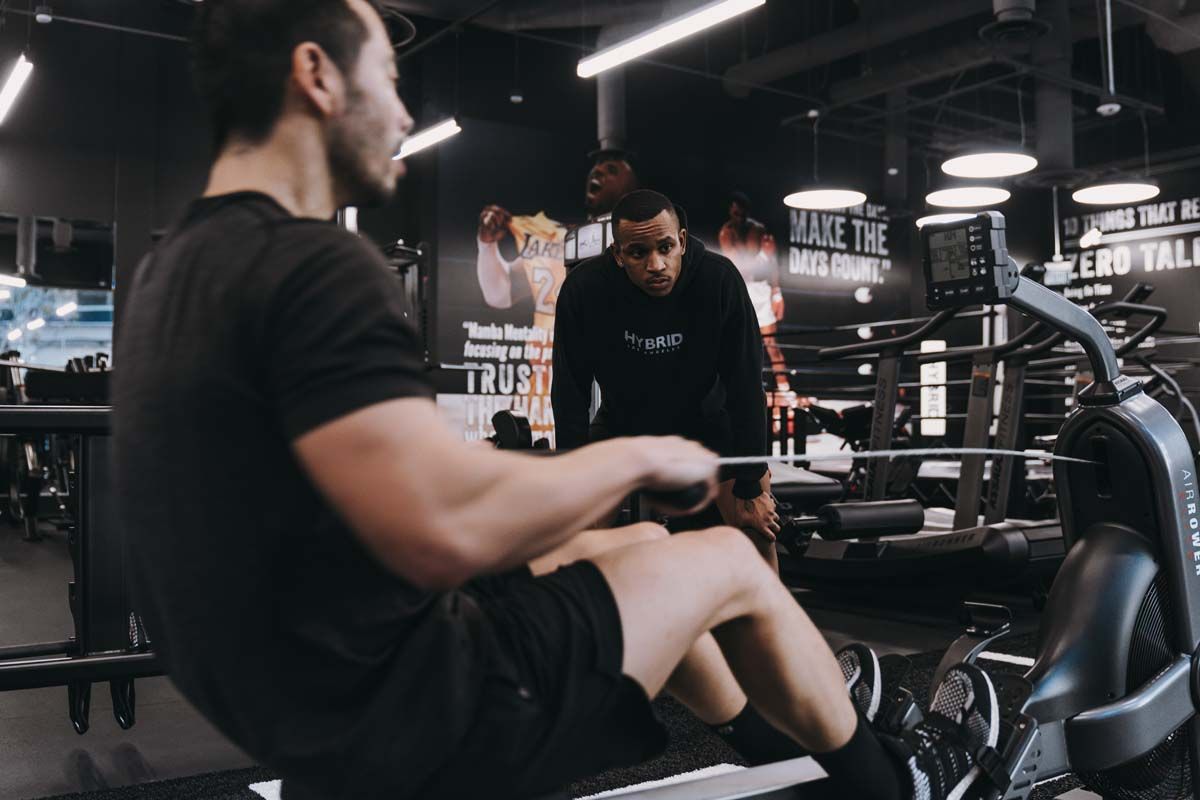 A man is riding a rowing machine in a gym while another man watches.