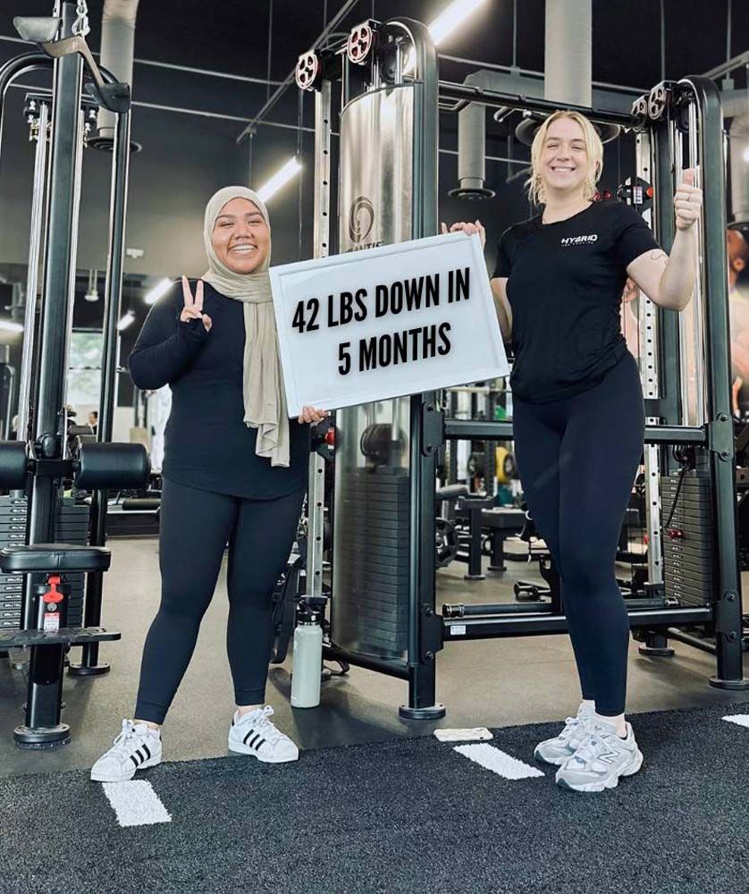 Two women are standing in a gym holding a sign that says 42 lbs down in 5 months
