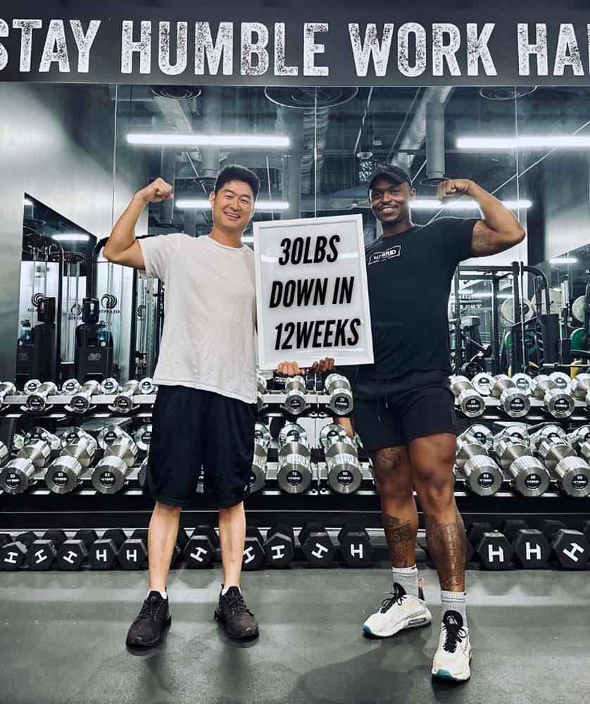 Two men are standing next to each other in a gym holding a sign that says 30lbs down in 12 weeks.