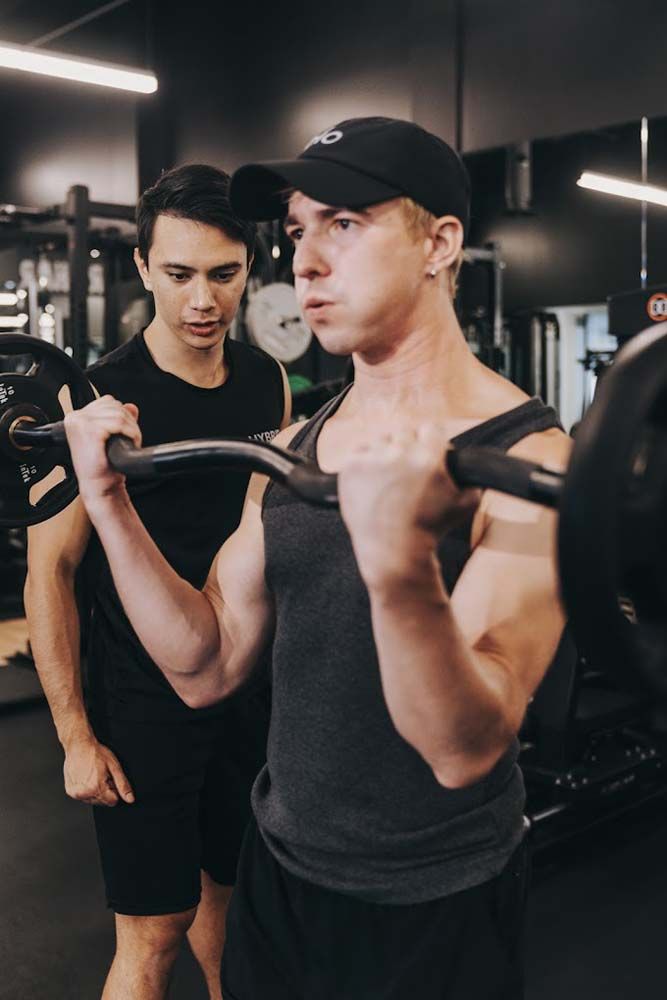 A man is lifting a barbell in a gym while another man watches.