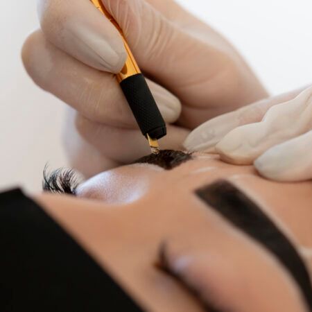 Close-up of a person receiving eyebrow microblading; a gloved hand holds a tool to apply ink to the brow.