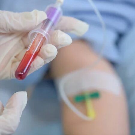 Gloved hand holding blood vial next to a patient’s arm with an IV needle inserted.