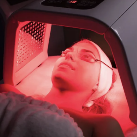 Woman in a red light therapy bed wearing eye protection and a white headband at a spa.