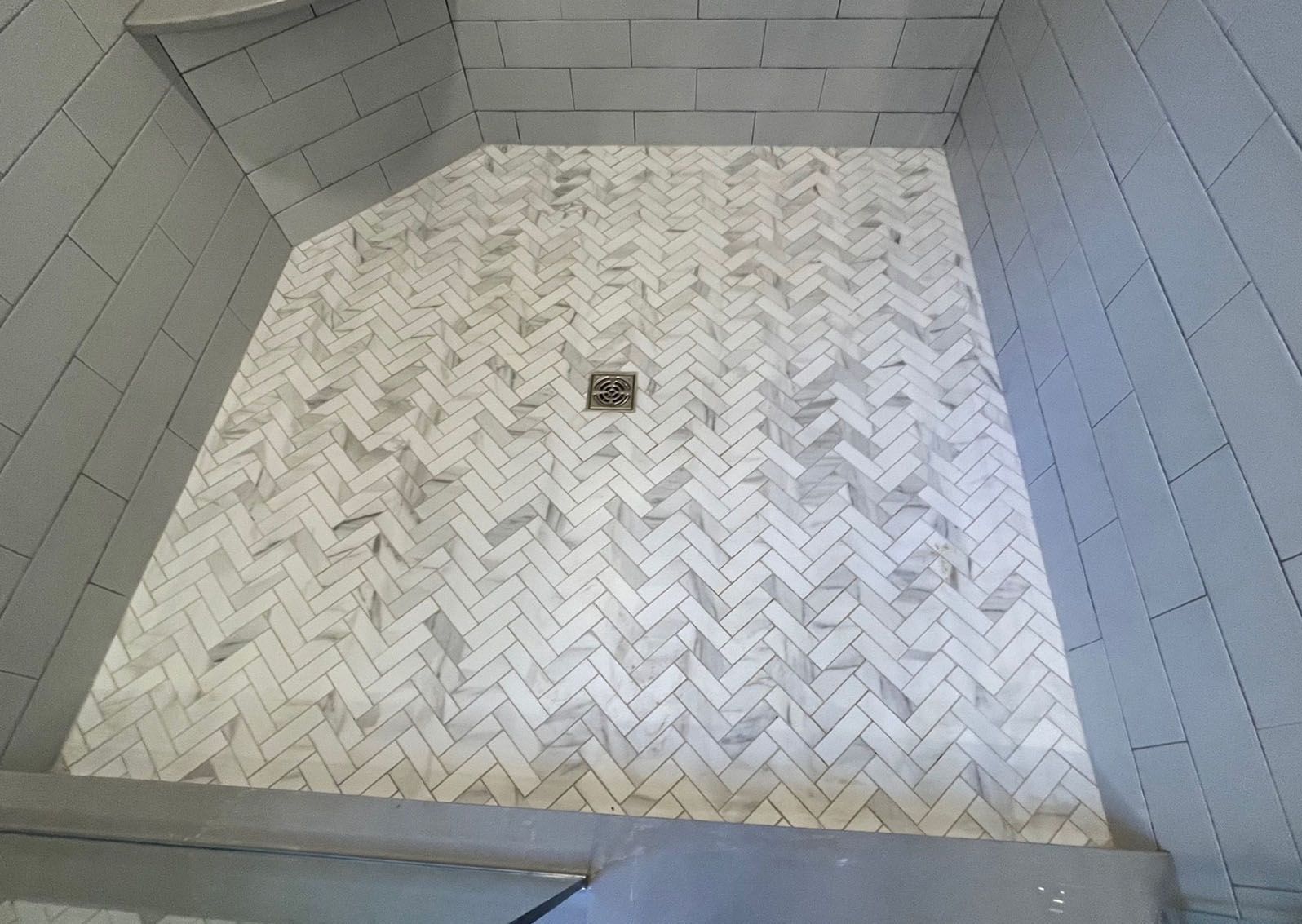 A shower stall with a herringbone tile floor and a drain.