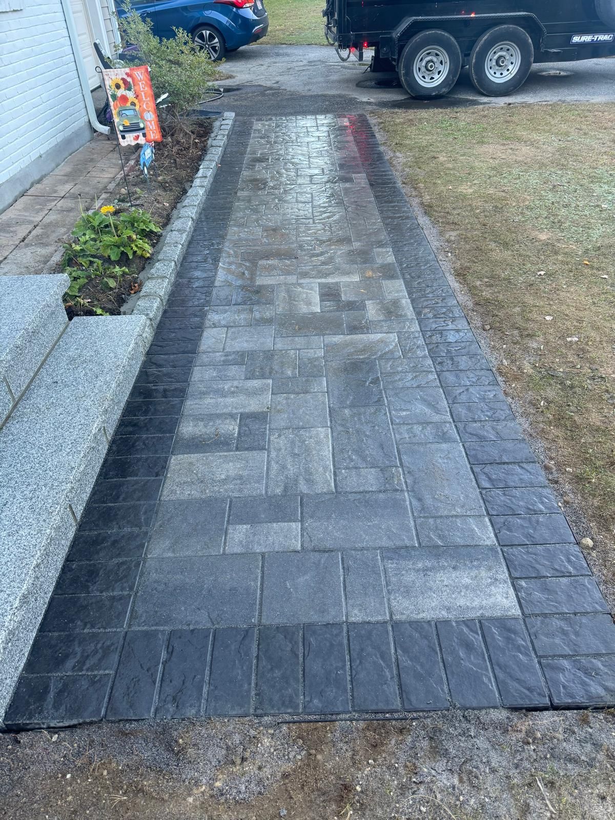A paved walkway made of gray rectangular stones bordered by dark charcoal bricks, leading from steps to a driveway.
