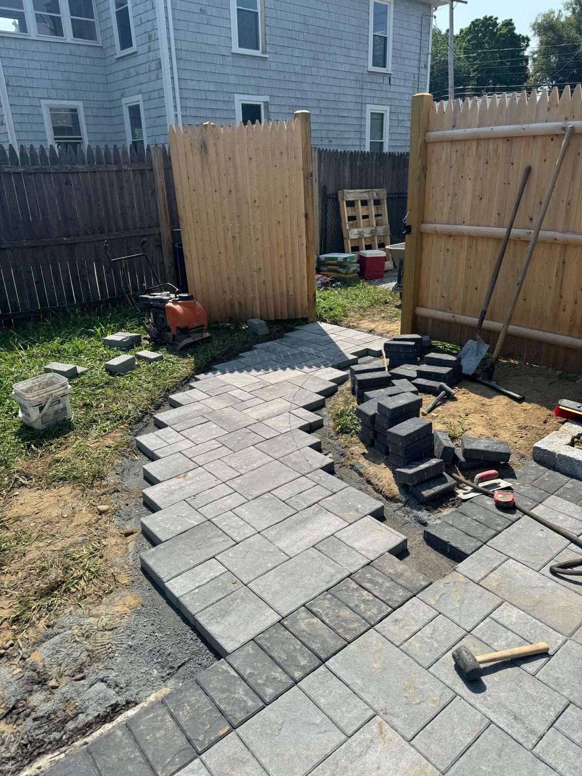 A patio under construction in a backyard, featuring a newly laid gray paver walkway, loose stones, and a wooden fence.