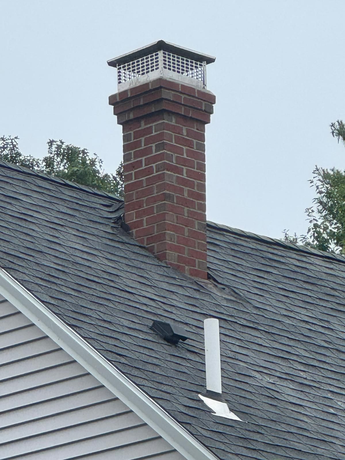 A red brick chimney with a metal cap sits on a grey shingled roof next to a white plumbing vent pipe.