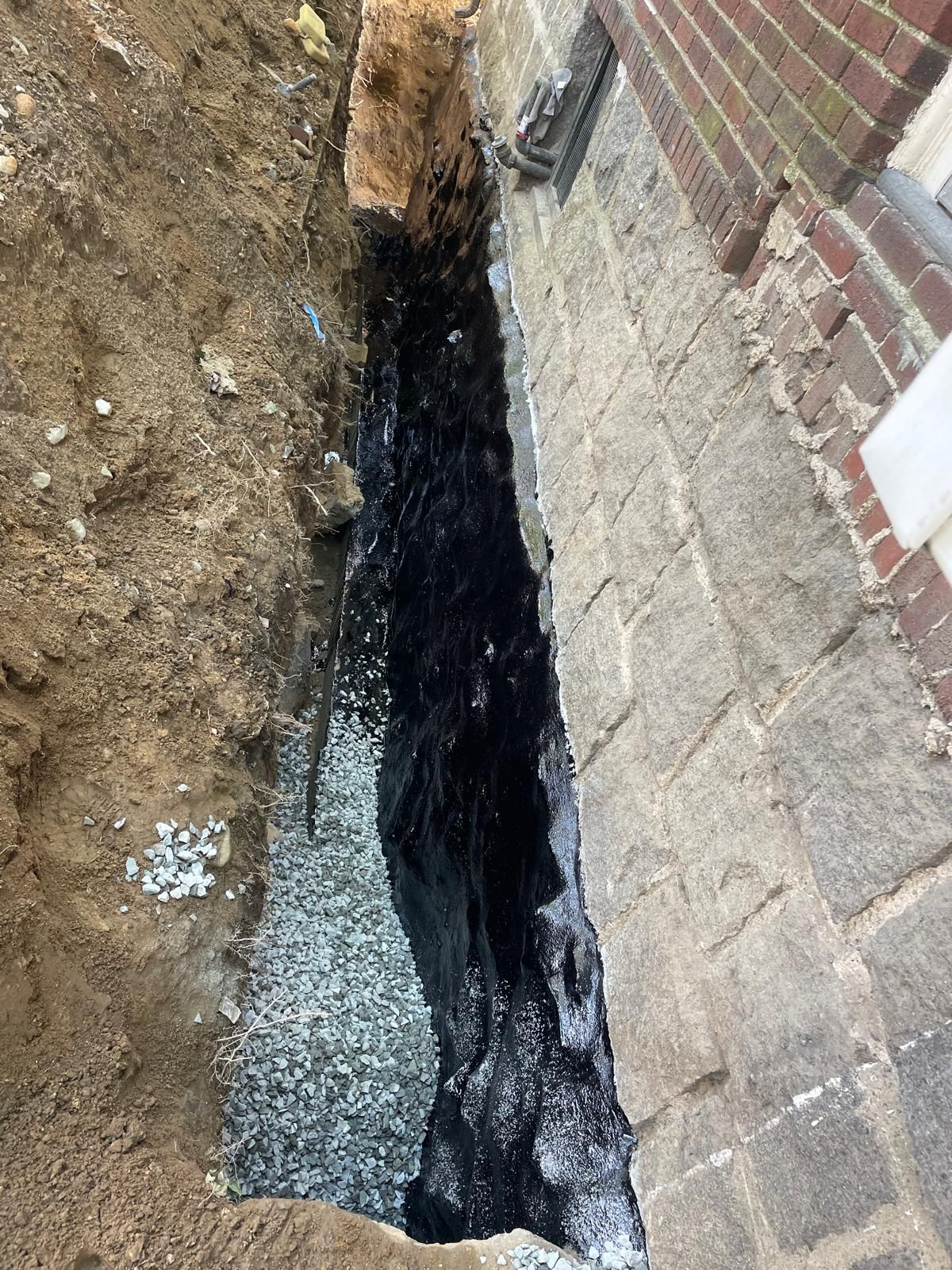 A trench dug against a stone house foundation shows a fresh, black waterproof coating applied over gray stone blocks.