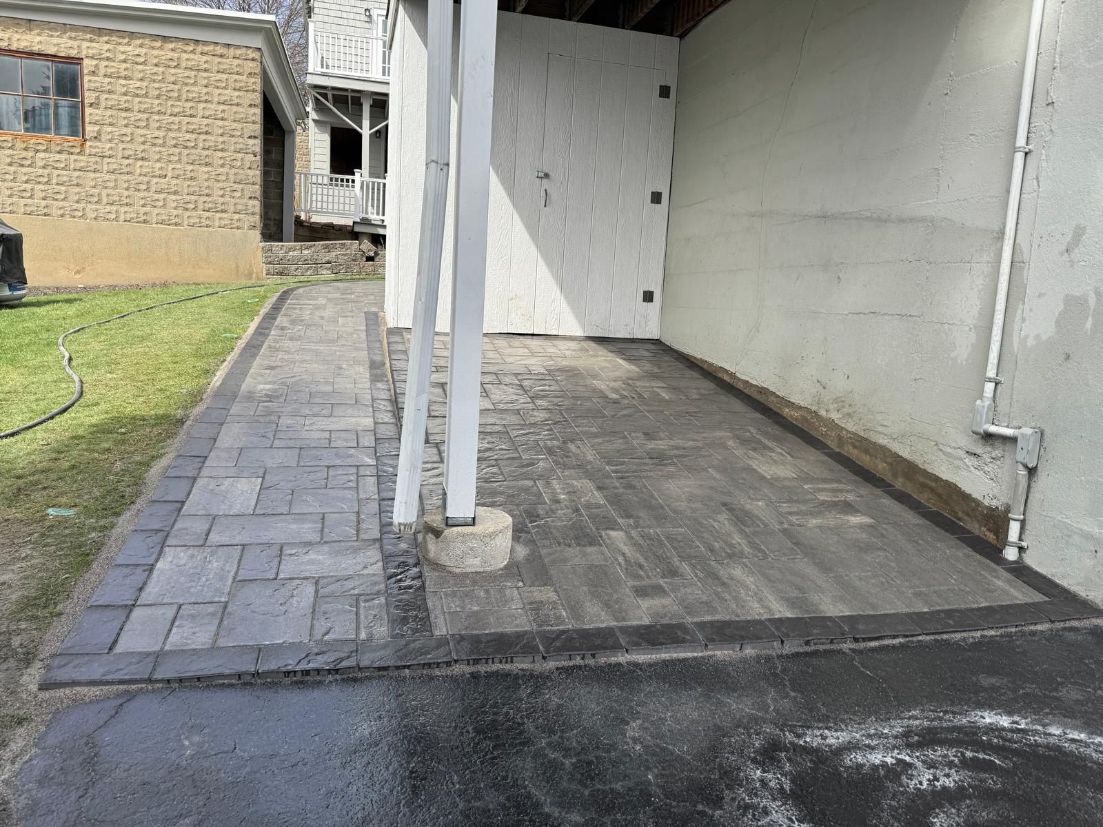 A paved walkway and patio area with gray rectangular stone pavers and a dark border, located next to a building wall.
