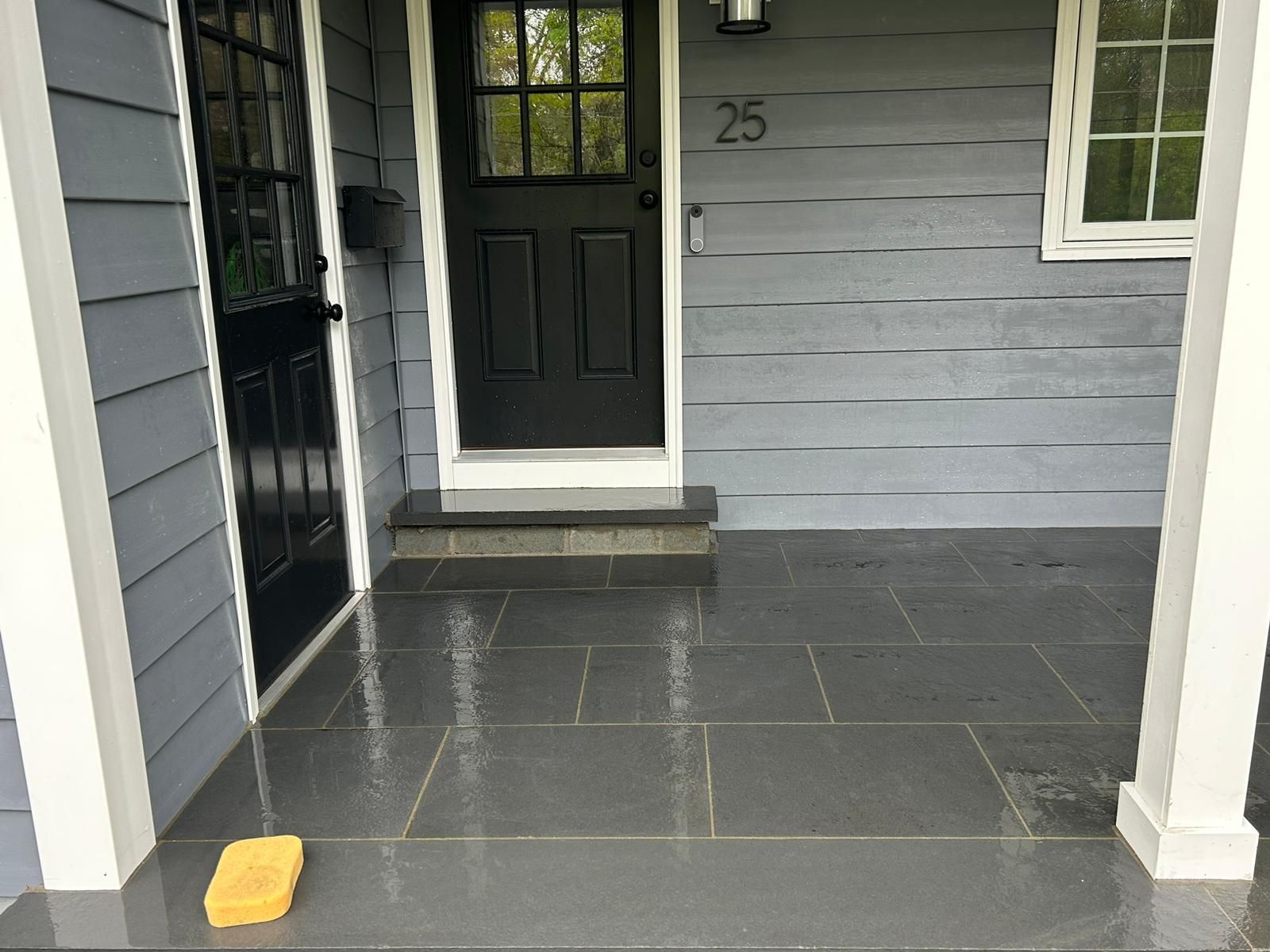 A front porch with grey stone tiles, blue siding, a black front door numbered 25, and a yellow sponge on the floor.