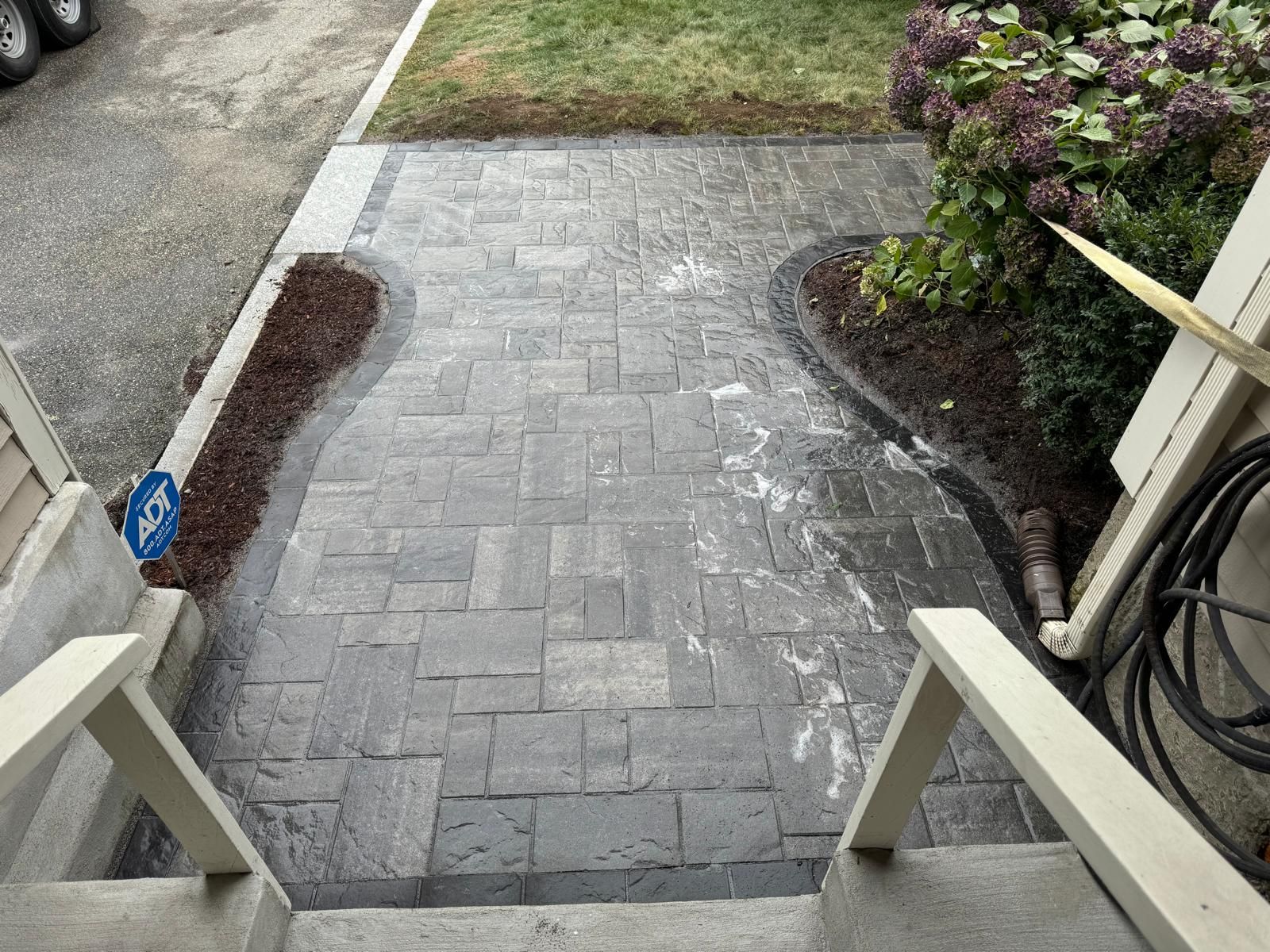 A stone paver walkway with a curved garden border leads from concrete steps toward a patch of grass.