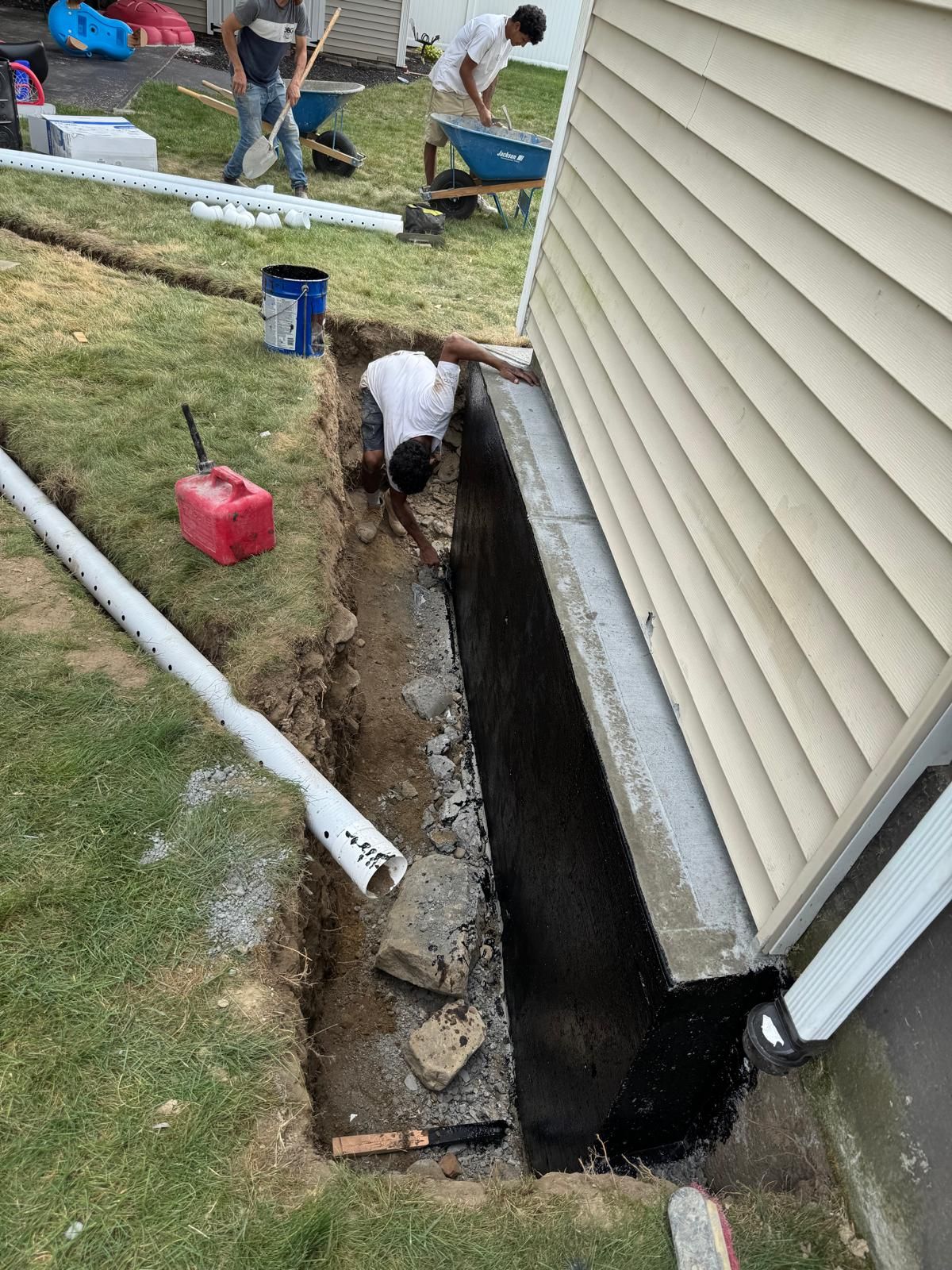 Workers excavate a trench alongside a house foundation coated in black sealant, with construction tools in the grass.