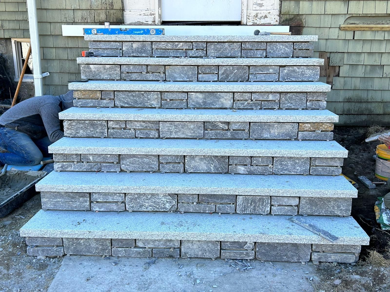 A worker installing a set of new stone stairs with light-colored granite treads and dark gray stacked stone risers.