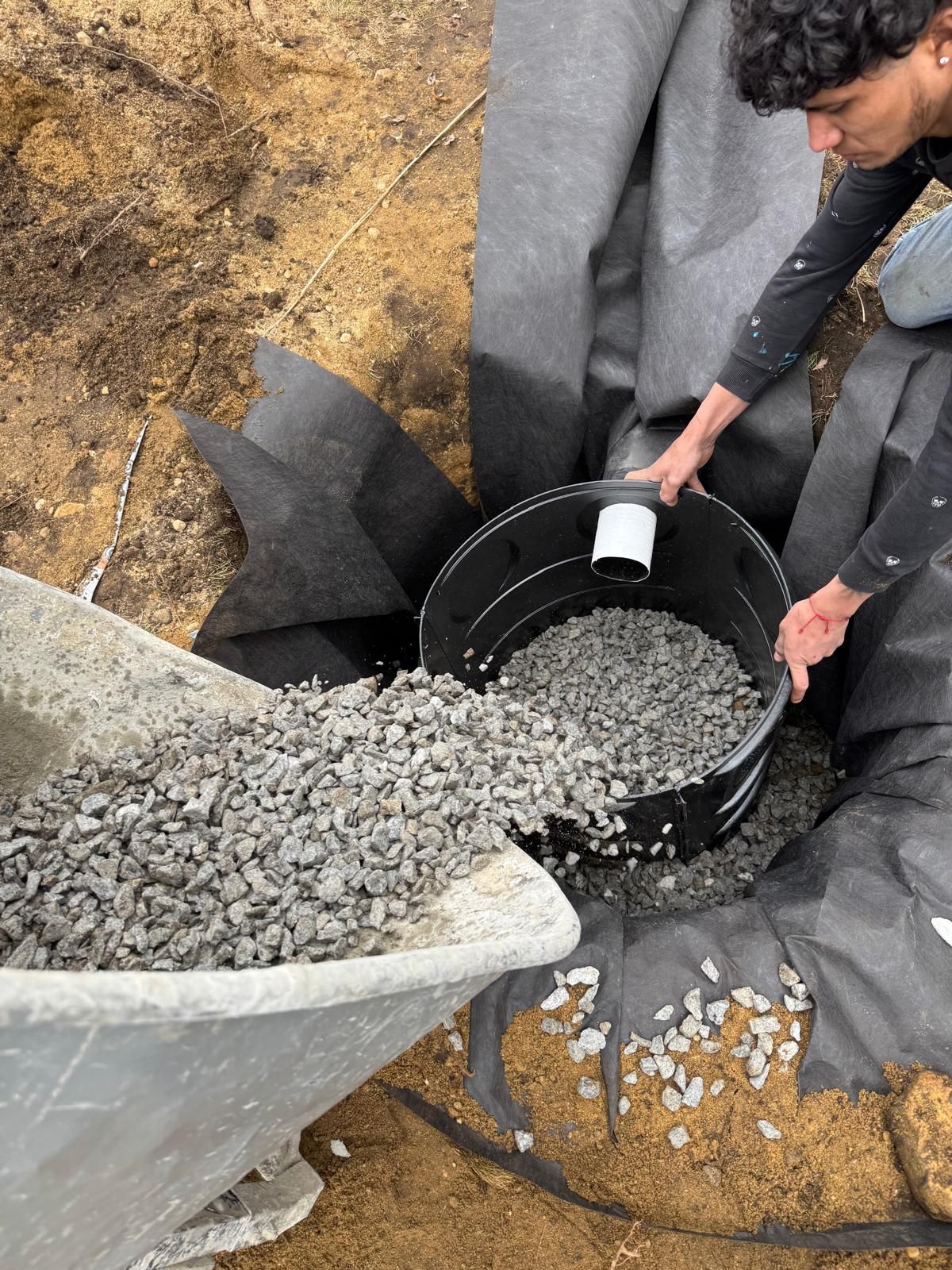 A person pouring gravel from a wheelbarrow into a black drainage basin lined with landscape fabric in a dirt pit.
